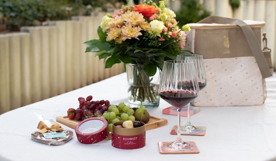 Ein Tisch, auf dem verschiedene Geschenkideen von Lebkuchen Schmidt stehen. Neben Pralinen, einem Schokoherz stehen auf dem Tisch Gläser mit Rotwein gefüllt, eine Kühltasche und eine Blumenvase mit einem bunten Blumenstrauß