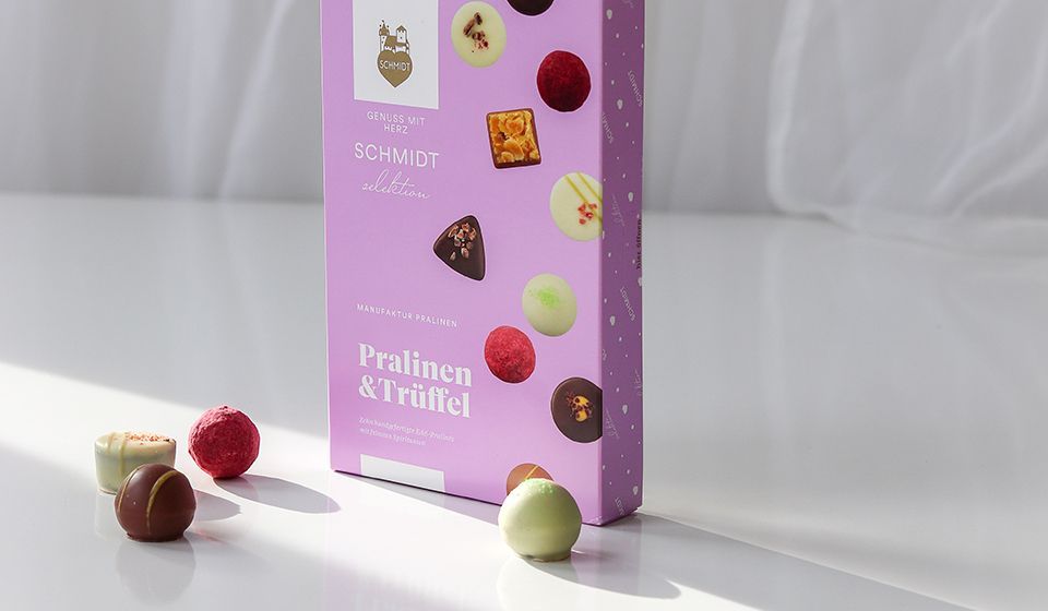 Produktfoto von Pralinen & Trüffel von Lebkuchen Schmidt, mit 4 Pralinen, die vor der Packung platziert sind. Im Hintergrund scheint die Sonne in den Raum